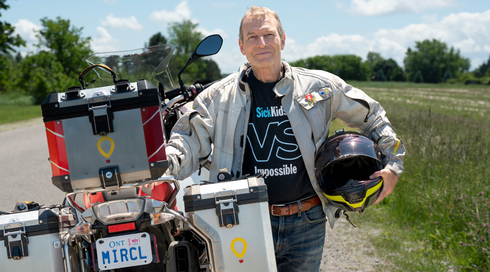 Bruce stands in a SickKidsVS shirt and a motorcycle jacket. He's leaning on his motorcycle which has a license plate that reads MRCL in honour of his Motorcycle for Miracles fundraising. -- Bruce porte une chemise SickKidsVS et une veste de moto. Il s'appuie sur sa moto dont la plaque d'immatriculation indique MRCL en l'honneur de sa collecte de fonds Motorcycle for Miracles.