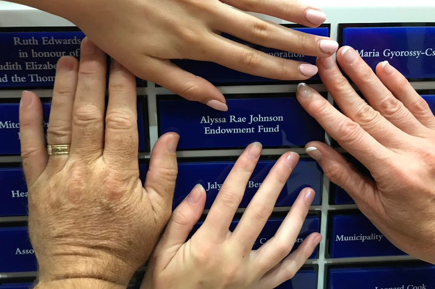 The hands of Bruce, Mary, Holly and Jocelyn can be seen encircling a plaque that reads: Alyssa Rae Johnson Endowment Fund -- On peut voir les mains de Bruce, Mary, Holly et Jocelyn encerclant une plaque qui dit : Alyssa Rae Johnson Endowment Fund