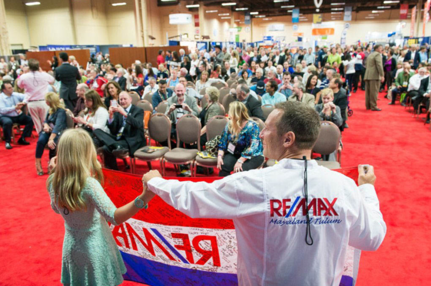 Holly and Bruce standing in front of a large crowd of REALTORS at a convention. They are holding one of the Motorcycle for Miracle flags that has been signed by brokerages from all over North America -- Holly et Bruce devant une grande foule d'agents immobiliers lors d'un congrès. Ils tiennent l'un des drapeaux Motorcycle for Miracle qui a été signé par des courtiers de toute l'Amérique du Nord